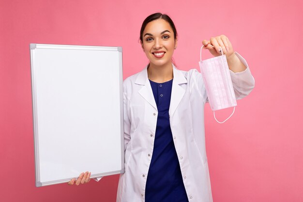 Femme médecin dans un manteau médical blanc tenant un tableau blanc avec espace de copie pour le texte et un masque de protection isolé