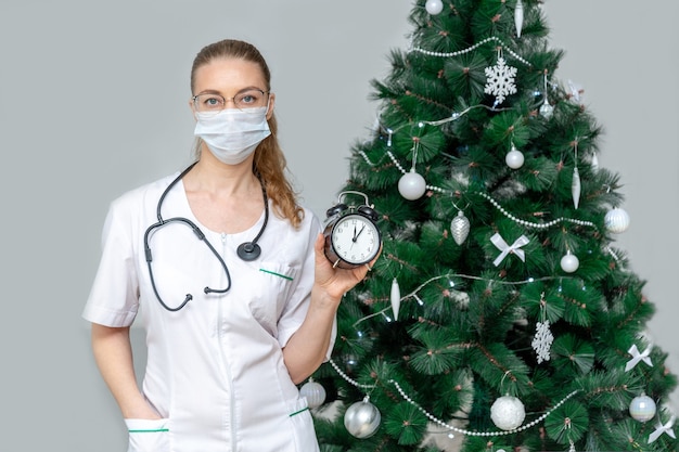 Femme médecin dans un masque médical de protection détient un réveil sur le fond d'un Noël