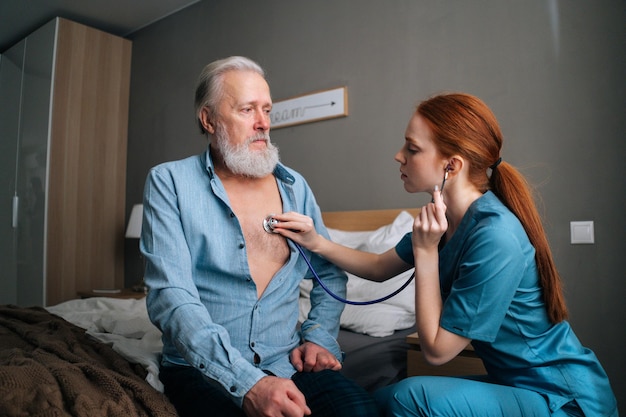 Femme médecin écoutant avec stéthoscope les poumons d'un patient âgé âgé assis sur son lit à la maison