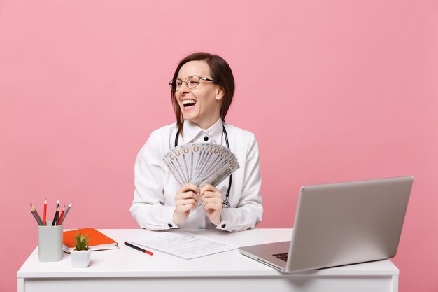 Une femme médecin est assise au bureau sur un ordinateur avec de l'argent en espèces pour un document médical à l'hôpital isolé sur fond de mur rose pastel. Femme en robe médicale lunettes stéthoscope. Concept de médecine de santé