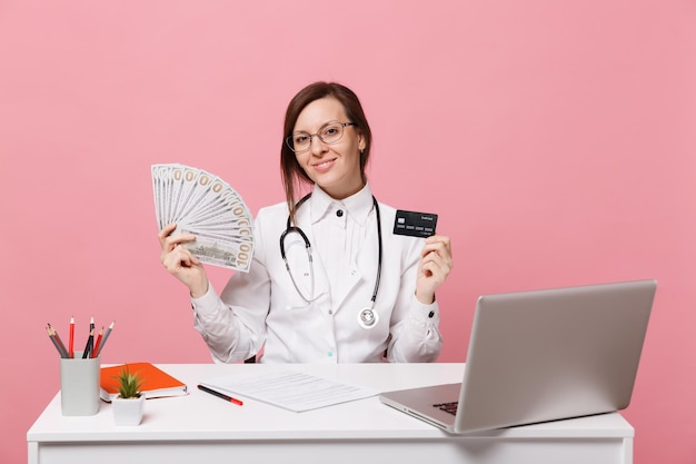 Une femme médecin est assise au bureau sur un ordinateur avec de l'argent en espèces pour un document médical à l'hôpital isolé sur fond de mur rose pastel. Femme en robe médicale lunettes stéthoscope. Concept de médecine de santé