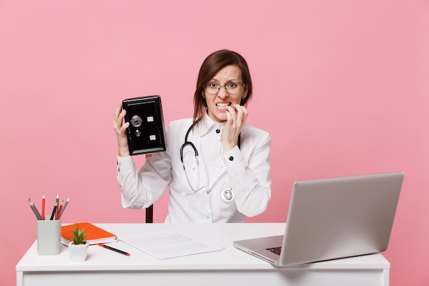 Une femme médecin est assise au bureau et travaille sur un ordinateur avec un document médical dans un coffre-fort à l'hôpital isolé sur fond rose pastel. Femme en robe médicale lunettes stéthoscope. Concept de médecine de santé.