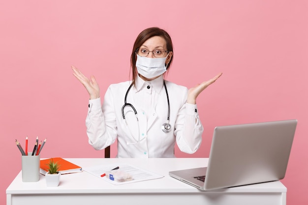 Une femme médecin est assise au bureau et travaille sur un ordinateur avec un document médical dans un masque facial à l'hôpital isolé sur fond de mur rose pastel. Femme en robe médicale lunettes stéthoscope Concept de médecine de santé