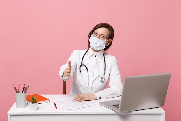 Une femme médecin est assise au bureau et travaille sur un ordinateur avec un document médical dans un masque facial à l'hôpital isolé sur fond de mur rose pastel. Femme en robe médicale lunettes stéthoscope Concept de médecine de santé