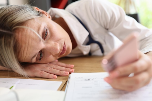 Femme médecin fatiguée allongée sur son bureau et regardant l'écran du smartphone