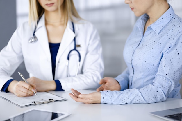 Une femme-médecin inconnue écoute son patient, assise ensemble au bureau dans l'armoire d'une clinique. Femme médecin avec un stéthoscope écrit au presse-papiers, gros plan. Médical parfait