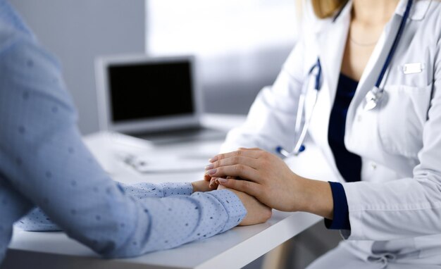 Une femme-médecin inconnue tient les mains de sa patiente, discutant de l'examen de santé actuel, alors qu'elle est assise au bureau dans l'armoire d'une clinique. Médecin avec un stéthscope au travail, gros plan