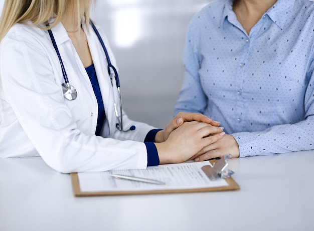 Une femme-médecin inconnue tient les mains de son patient pour rassurer un patient, alors qu'elle est assise ensemble au bureau dans l'armoire d'une clinique. Femme médecin utilise un presse-papiers et un stéthoscope, à proximité