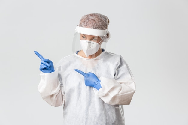 Femme médecin intriguée et réfléchie en équipement de protection individuelle, écran facial et gants, regardant et pointant le coin supérieur gauche.