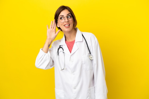 Femme médecin isolée sur fond jaune écoutant quelque chose en mettant la main sur l'oreille