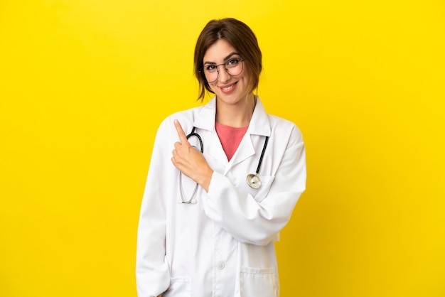 Femme médecin isolée sur fond jaune pointant vers le côté pour présenter un produit