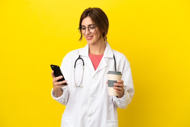 Femme médecin isolée sur fond jaune tenant du café à emporter et un mobile