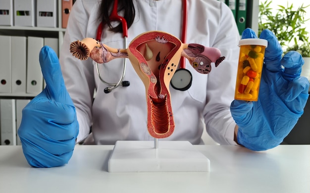 Photo une femme médecin joyeuse en gants bleus tenant un modèle d'utérus et une bouteille de pilules montrant le pouce en l'air.