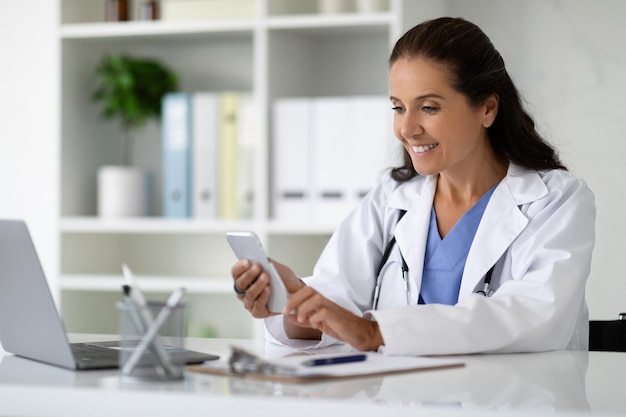 Femme médecin joyeuse et mature assise dans son cabinet à l'aide de gadgets