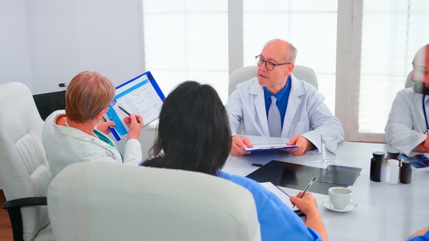 Femme médecin mature pointant sur le presse-papiers pendant le briefing avec des collègues travaillant dans la salle de réunion de l'hôpital. Thérapeute expert de la clinique discutant avec des collègues de la maladie, professionnel de la médecine