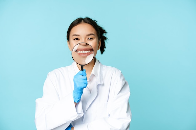 Femme médecin médecin asiatique drôle montrant son sourire avec une loupe debout dans un unif médical...