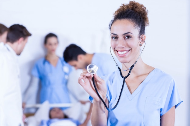 Femme médecin montrant un stéthoscope en direction de la caméra et autre médecin examinant un patient à l&#39;hôpital