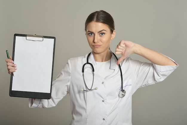 Femme médecin montre presse-papiers vide vide et pouce vers le bas sur fond gris