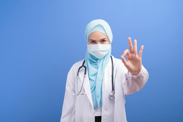 Une femme médecin musulmane avec hijab portant un masque chirurgical sur fond bleu studio.