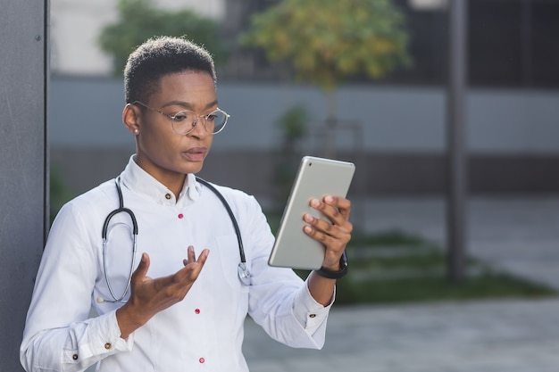 Une femme médecin organise une consultation en ligne via un lien vidéo tenant une tablette, près de la clinique, une conférence à l'heure du déjeuner avec le personnel