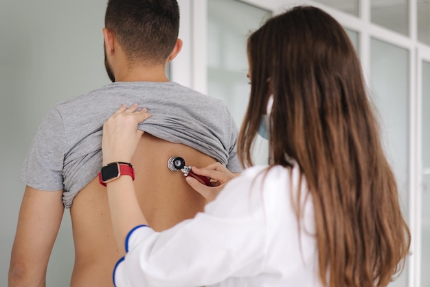 Femme médecin portant un masque médical à l'aide d'un stéthoscope pour vérifier les poumons d'un patient en clinique médicale