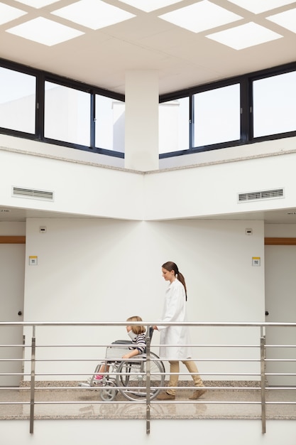 Femme médecin poussant l&#39;enfant avec une minerve en fauteuil roulant