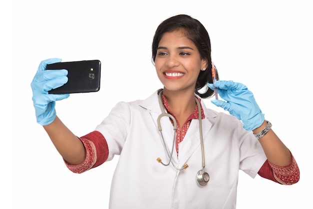 Femme médecin prenant selfie avec stéthoscope et injection par smartphone sur fond blanc.