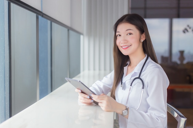 Une femme médecin professionnelle asiatique vêtue d'une blouse médicale blanche est assise sur une chaise dans une bibliothèque d'hôpital pour rechercher des informations sur une tablette pour un traitement dans le concept de soins de santé.