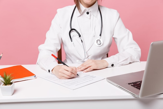 Femme médecin recadrée assise au bureau travaillant à remplir des documents médicaux à l'hôpital isolé sur fond de mur rose pastel à l'intérieur. Femme en stéthoscope de robe médicale. Concept de médecine de santé.