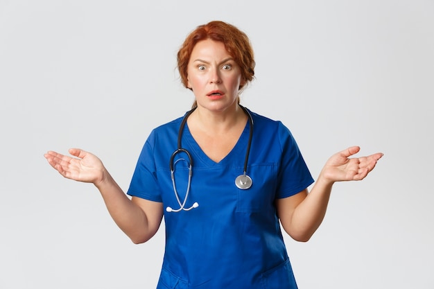 Femme médecin rousse inquiète et confuse, l'infirmière ne peut pas comprendre ce qui s'est passé, portant des gommages bleus, écartant les mains sur le côté et semblant inquiète