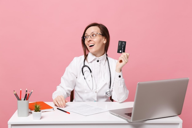 Femme médecin s'asseoir au bureau travailler sur ordinateur avec carte de crédit de document médical à l'hôpital isolé sur fond de mur rose pastel. Femme en robe médicale lunettes stéthoscope. Concept de médecine de santé