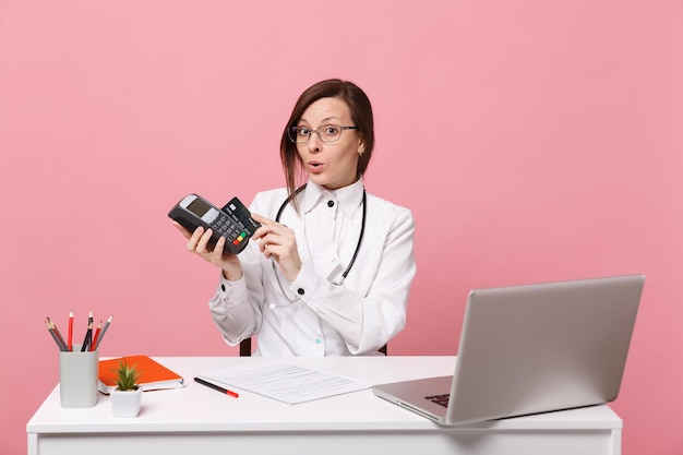 Femme médecin s'asseoir au bureau travailler sur ordinateur avec carte de crédit de document médical à l'hôpital isolé sur fond de mur rose pastel. Femme en robe médicale lunettes stéthoscope. Concept de médecine de santé