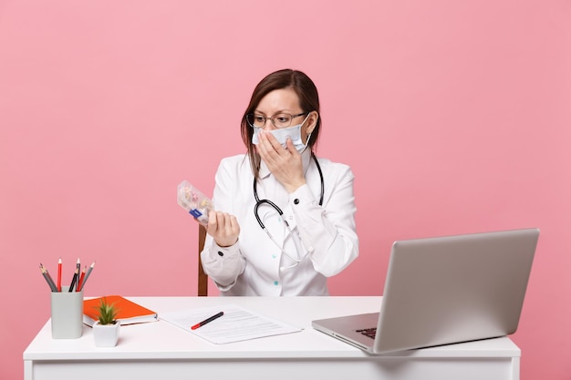 Femme médecin s'asseoir au bureau travailler sur ordinateur avec document médical tenir des pilules à l'hôpital isolé sur fond de mur rose pastel. Femme en robe médicale lunettes stéthoscope. Concept de médecine de santé.