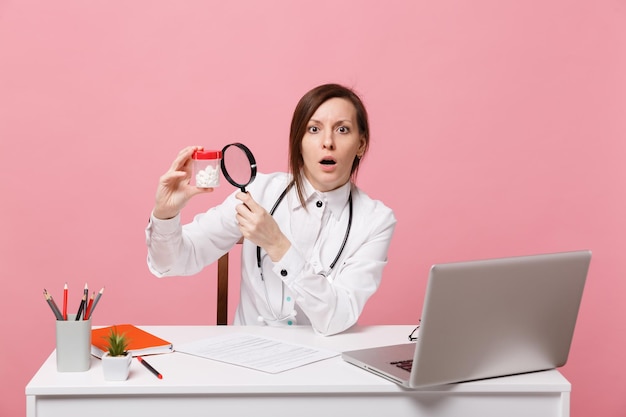 Femme médecin s'asseoir au bureau travailler sur ordinateur avec document médical tenir des pilules à l'hôpital isolé sur fond de mur rose pastel. Femme en robe médicale lunettes stéthoscope. Concept de médecine de santé.