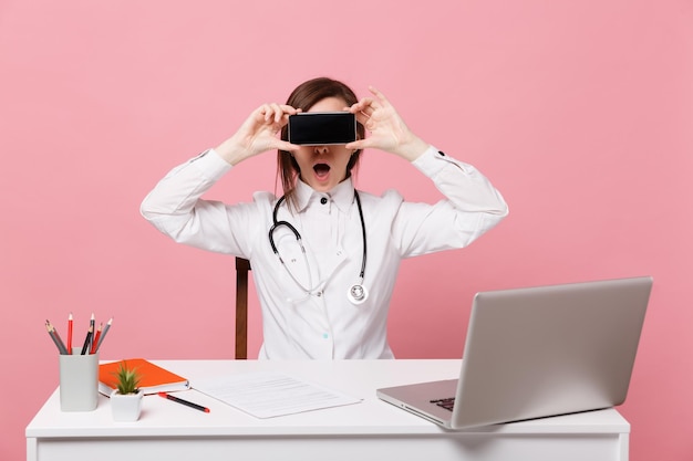 Femme médecin s'asseoir au bureau travailler sur ordinateur avec document médical tenir téléphone portable à l'hôpital isolé sur fond rose pastel. Femme en robe médicale lunettes stéthoscope. Concept de médecine de santé.