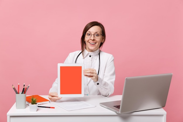 Une femme médecin s'assoit au bureau sur un ordinateur avec une tablette de maintien de document médical à l'hôpital isolée sur fond de mur rose pastel. Femme en robe médicale lunettes stéthoscope. Concept de médecine de santé