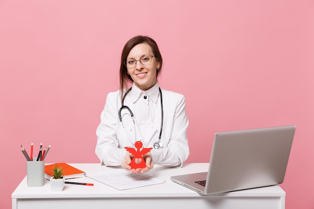 Une femme médecin s'assoit au bureau et travaille sur un ordinateur avec un document médical tenant un ange à l'hôpital isolé sur fond de mur rose pastel. Femme en robe médicale lunettes stéthoscope. Concept de médecine de santé.