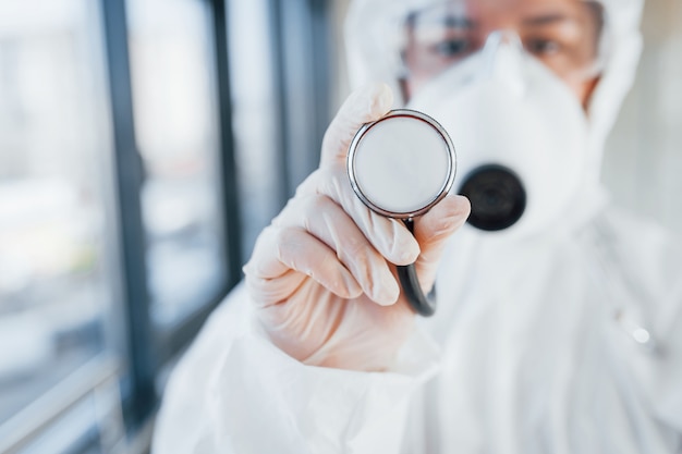 Femme médecin scientifique en blouse de laboratoire, lunettes défensives et masque debout à l'intérieur et détient un stéthoscope