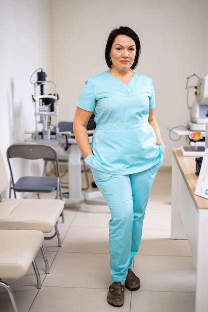 Femme médecin se tient les bras dans les poches et regarde la caméra Clinique moderne avec équipement à jour pour les tests oculaires Gros plan