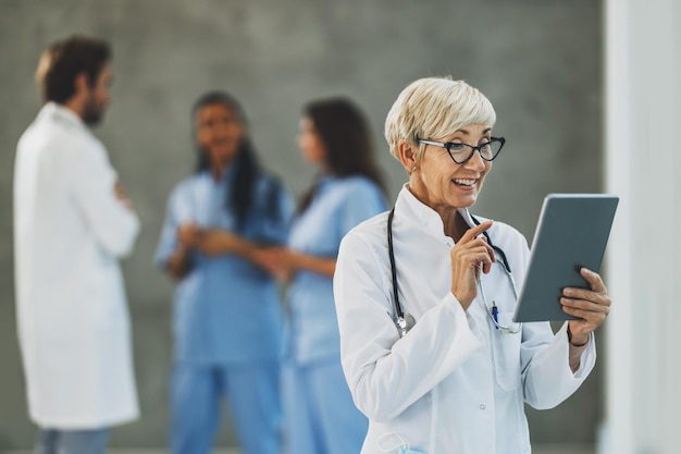 Une femme médecin senior qui a réussi à passer un appel vidéo sur une tablette numérique pendant une pause rapide dans un couloir d'hôpital.