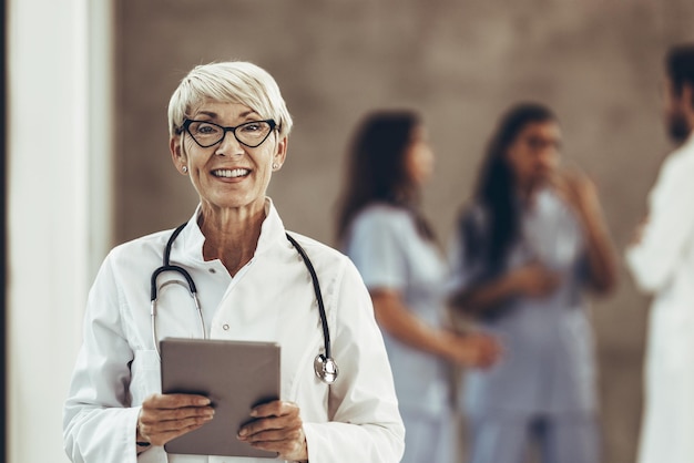 Une femme médecin senior réussie regardant la caméra et tenant une tablette numérique dans un couloir d'hôpital.