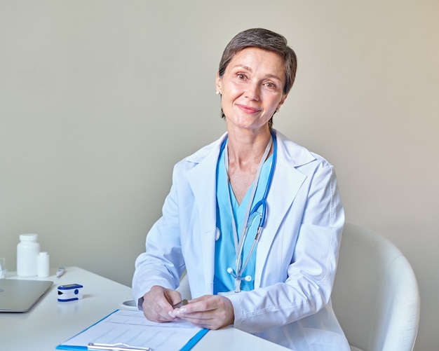 Femme médecin souriante d'âge moyen avec stéthoscope sur les épaules en blouse blanche travaillant en clinique