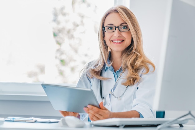 Femme médecin souriante assise à son bureau tenant une tablette numérique