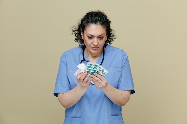 femme médecin suspecte d'âge moyen portant un uniforme et un stéthoscope autour du cou tenant des paquets de pilules avec les deux mains les regardant isolées sur fond olive