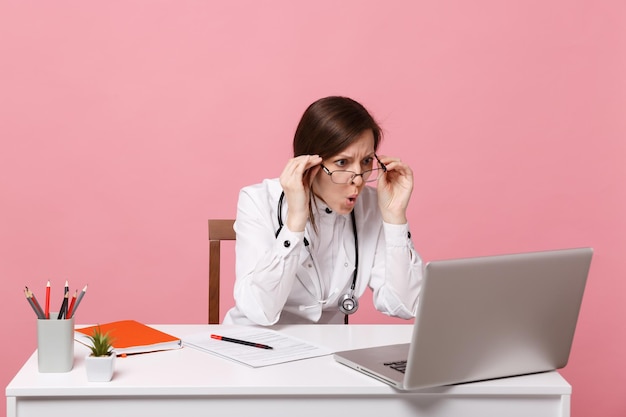 Une femme médecin triste et fatiguée est assise au bureau sur un ordinateur avec un document médical à l'hôpital isolé sur fond de mur rose pastel. Femme en robe médicale lunettes stéthoscope. Concept de médecine de santé.