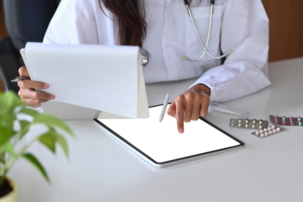 Une femme médecin en uniforme blanc tenant des documents et vérifiant des informations médicales sur une tablette numérique.