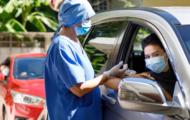 Une femme médecin en uniforme d'hôpital bleu et un masque facial se tiennent près d'une voiture tenant une seringue dans les mains et portent des gants en caoutchouc injectant le vaccin covid 19 pour traverser l'épaule du patient dans la file d'attente de vaccination.