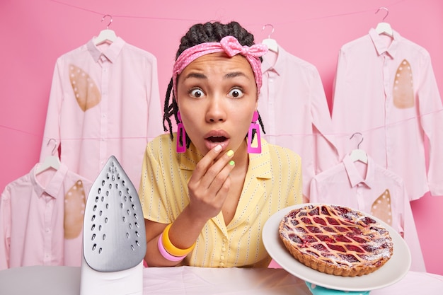Femme de ménage abasourdie à la peau foncée avec des dreadlocks regarde la caméra surprise en train de cuisiner une tarte savoureuse occupée à repasser à la maison habillée avec désinvolture isolée sur un mur rose