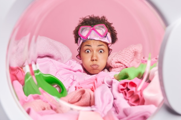 Photo femme de ménage afro-américaine souffle les joues fait la grimace a une expression surprise porte un masque de plongée en apnée sur le front entouré de linge sale et de poses de détergent à l'intérieur de la machine à laver