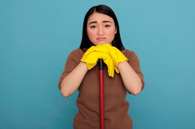 Femme de ménage asiatique accablée, triste et fatiguée, prenant du repos portant des gants jaunes tenant un bâton de brosse isolé sur fond bleu, concept de maison de nettoyage, femme surmenée se sentant négative